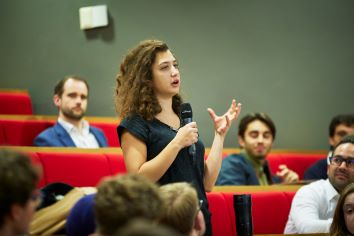 Young female holding mic, standing in theatre, asking a question, gesticulating animatedly