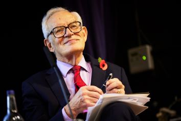 Charles Goodart smiling while chairing Andrea Enria lecture in October 2023 at LSE, wearing a dark suit with pink shirt, red tie, and a poppy on the lapel, holding a pen