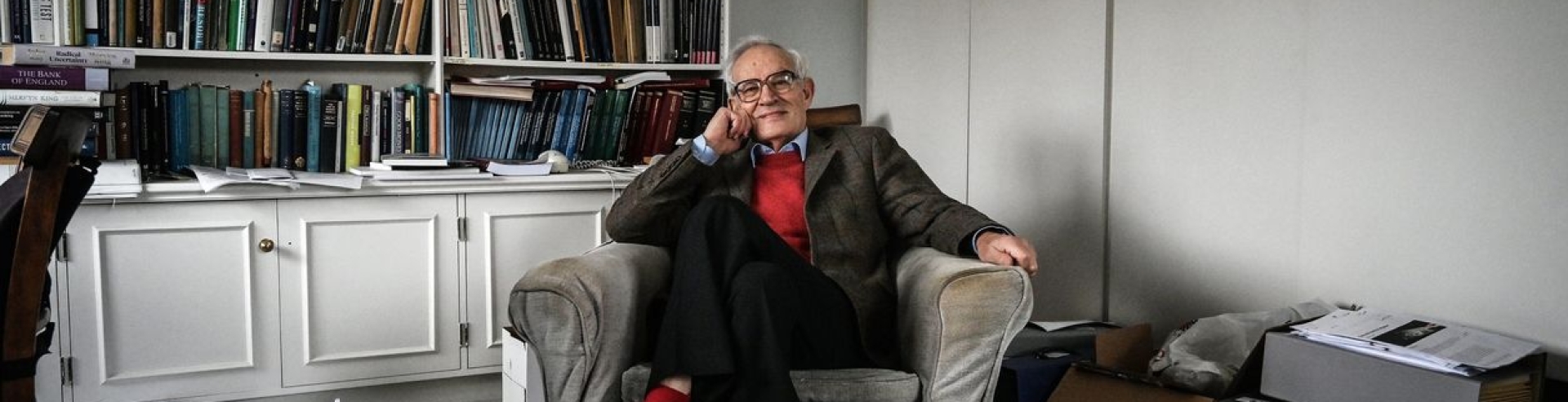 Charles Goodhart in his home, sat in an armchairm, surrounded by his books