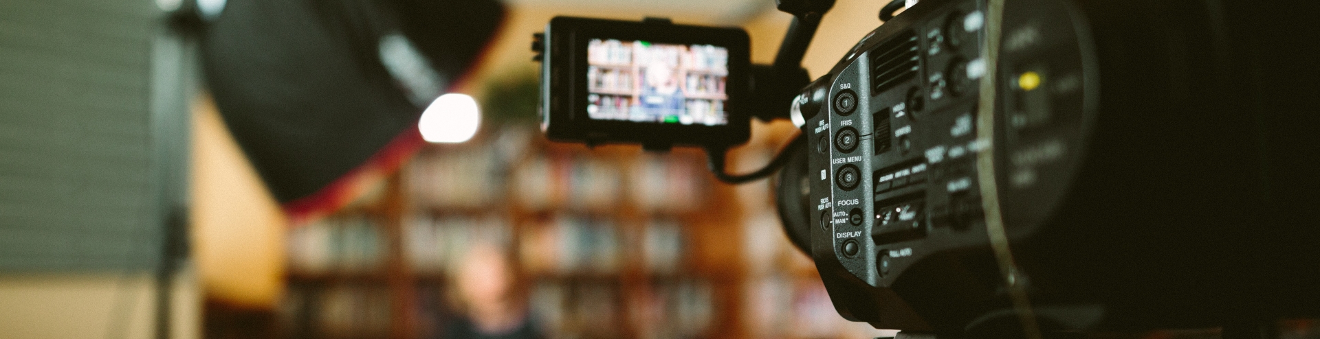 Camera in focus  and in background a blurred image of a female interviewee sat behind a bookcase, in an office