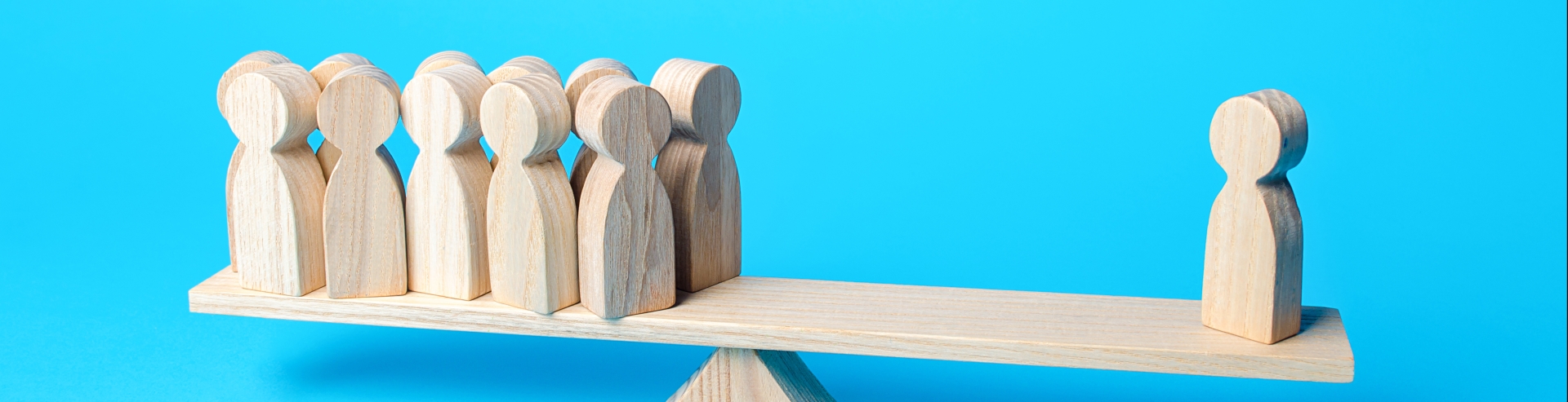 Human figurine peg balancing on a see saw. A group of human figurines on the other side. The see saw is in balance, suggesting the one has equal power, thus weight, compared to the group.